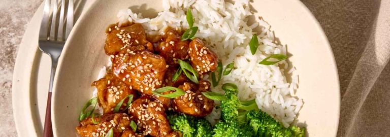 Close-up of sesame chicken coated in a sticky soy garlic sauce, served with steamed broccoli and topped with green onions and sesame seeds.