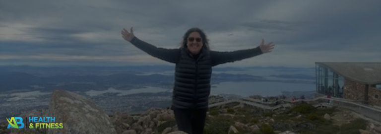 Woman in sunglasses and black jacket sitting on rocks with a scenic view of a coastal city and hills in the background.