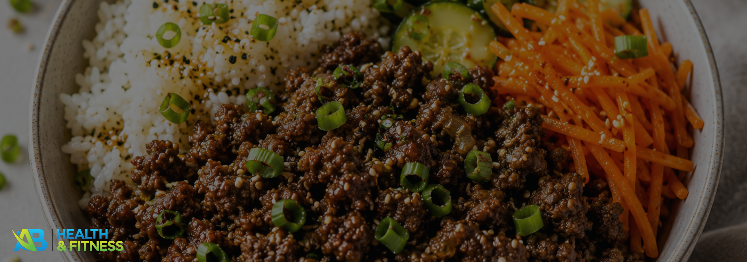 Close-up view of a savory Korean beef rice bowl garnished with fresh green onions and toasted sesame seeds next to vibrant green veggies.